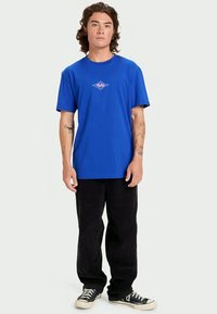 Μπλε βαμβακερό t-shirt με κοντά μανίκια και ένα μικρό μοβ και τυρκουάζ γραφικό λογότυπο στο στήθος. Συνδυάζεται με μαύρο παντελόνι και αθλητικά παπούτσια.