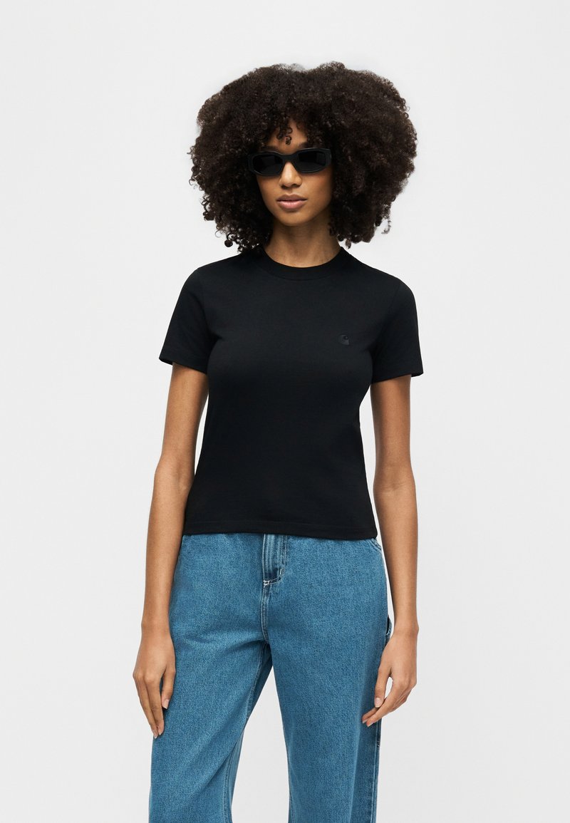 Woman with curly hair wearing black sunglasses, a black fitted T-shirt, and blue high-waist jeans, standing against a plain background.