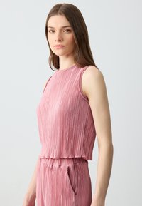 Top plissettato in morbido tessuto rosa, con scollo rotondo e design senza maniche. La texture è liscia con delicate linee verticali.