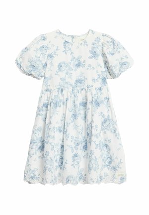 Uśmiechnięta młoda dziewczyna w białej sukience z niebieskimi kwiatowymi wzorami, beżowych butach i niebieskiej wsuwce do włosów, stojąca z rękami za plecami.