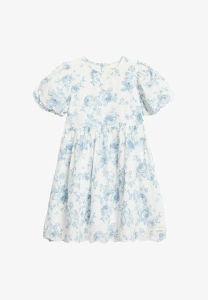 Lächelndes junges Mädchen in einem weißen Kleid mit blauen Blumenmustern, beigen Schuhen und einer blauen Haarklammer, steht mit den Händen hinter dem Rücken.