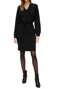 Kobieta w czarnej, długiej sukience do kolan z dekoltem w kształcie litery V i wiązanym paskiem w talii, w połączeniu z czarnymi przezroczystymi rajstopami i czarnymi botkami za kostkę.