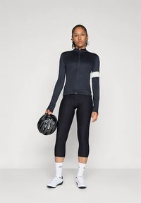 Chaqueta de ciclismo negra con cuello alto y cremallera, combinada con mallas ajustadas negras. El modelo sostiene un casco negro y lleva calcetines blancos y zapatillas de ciclismo.