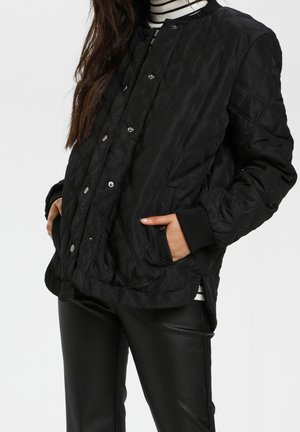 Vrouw draagt een zwarte gewatteerde jas met drukknopen en handen in de zakken, gecombineerd met zwarte leren broek en een gestreepte coltrui.