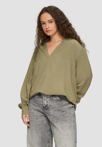 Olijfgroene blouse met lange mouwen, een V-hals en geassembleerde manchetten, gecombineerd met grijze denim jeans met een vervaagde textuur.