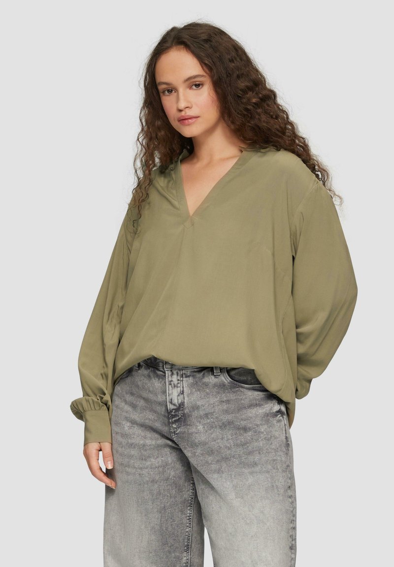 Olijfgroene blouse met lange mouwen, een V-hals en geassembleerde manchetten, gecombineerd met grijze denim jeans met een vervaagde textuur.