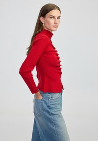 Czerwona bluzka z długim rękawem i guziczkami z przodu, wysoki kołnierz oraz dopasowany krój, w zestawie z jasnoniebieskimi dżinsami.