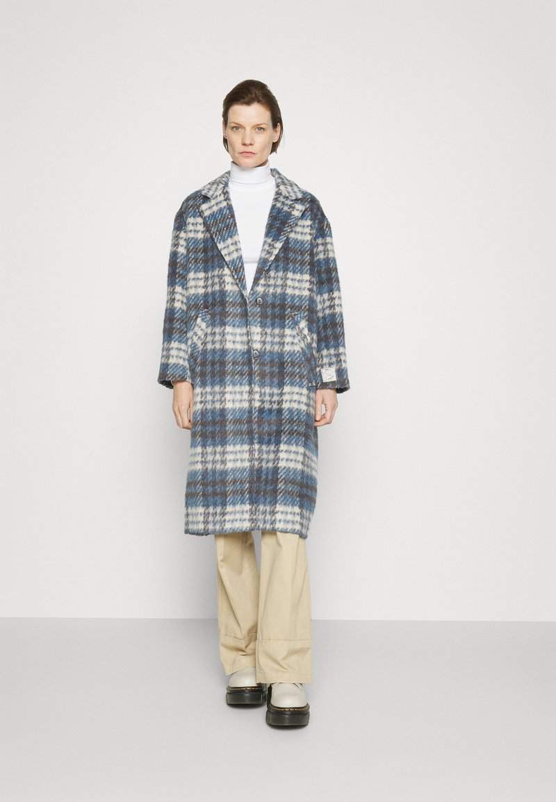 Abrigo de lana a cuadros en azul, blanco y gris; presenta un cuello con muesca, dos botones y bolsillos laterales; combinado con pantalones beige y zapatos blancos.