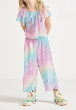 Enfant portant un haut plissé pastel arc-en-ciel et un pantalon avec des baskets montantes iridescentes, se tenant avec les mains derrière le dos.