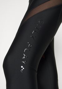 Leggings atléticas pretas com um tecido suave e detalhes em malha nas laterais. Um logo refletor está exibido na parte inferior da perna.