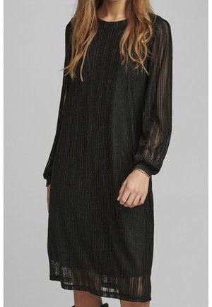 Femme portant une robe noire longueur genoux avec des rayures verticales scintillantes et des manches longues transparentes, accessoirisée d’un délicat bracelet en or.
