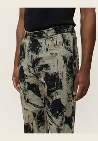 Green and black patterned trousers made of textured fabric, featuring a relaxed fit, side pockets, and a button closure.