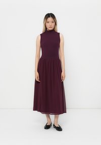 Woman standing against white background wearing sleeveless dark purple midi dress and black ballet flats.