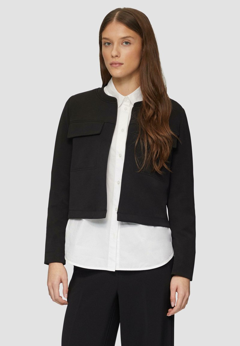 Schwarze, taillierte Jacke mit rundem Kragen, vorderen Taschen und langen Ärmeln, getragen über einem weißen Hemd mit Knöpfen. Glattes, strukturiertes Material.