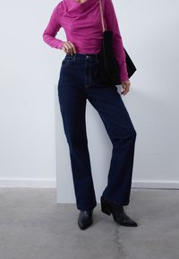 Hochgeschnittene, dunkelblaue Jeans aus Denim, kombiniert mit einem figurbetonten, langen, rosa Oberteil und schwarzen, hochhackigen Stiefeletten. Strukturierter Stoff und geformte Silhouette.