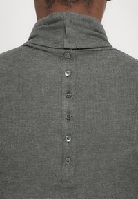 Man in een grijze ribbeltrui met een verticale rij van zes knopen langs de bovenrug onder een gevouwen hoge kraag.