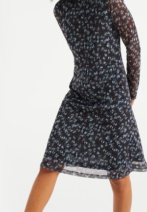 Kobieta w ciemnej, półprzezroczystej sukience z długim rękawem, ozdobionej niebiesko-białym kwiatowym wzorem, stoi rozkładając nogi na białym tle.