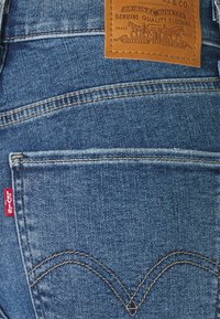 Vaqueros de mezclilla azul con un parche de cuero con el nombre de la marca, un bolsillo trasero con detalle de costura y una etiqueta roja en el lateral.