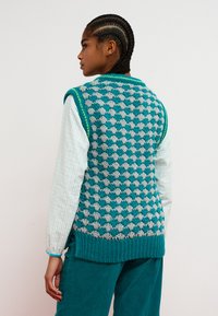 Vestă tricotată cu model de inimă turcoaz și gri, având tiv striat și margine verde. Poartă peste o cămașă cu mâneci lungi în carouri deschise.