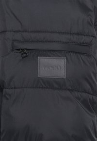 Black padded jacket featuring a zippered pocket on the chest and a rectangular logo patch. Smooth texture and minimalist design.