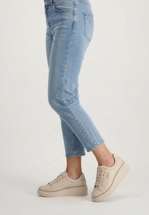 Beine in hellblauen, verkürzten Jeans und beigen Plateau-Sneakern mit der Aufschrift „VENEZIA“ an der Seite, posieren vor neutralem Hintergrund.