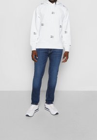 Witte capuchontrui met zwarte logo's, trekkoorden en geribbelde manchetten, gecombineerd met blauwe slim-fit jeans en witte sneakers met marineblauwe accenten.