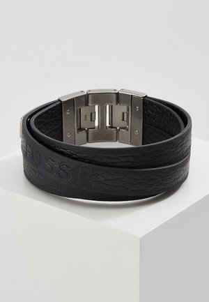 Czarna skórzana bransoletka typu wrap z fakturą, z wytłoczonym napisem i szerokim metalowym zapięciem, prezentowana na białej podstawce.