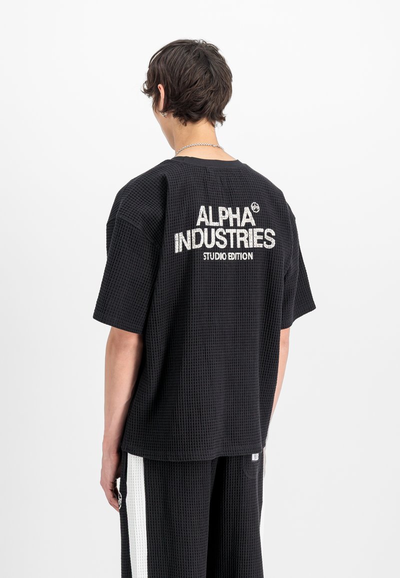 Person iført svart kortermet skjorte med vaffelmønster og bukser med hvite striper, skjorten har teksten "ALPHA INDUSTRIES STUDIO EDITION" på ryggen.