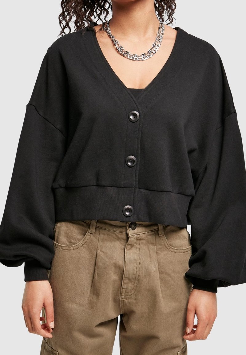 Femme porte un cardigan noir court boutonné avec des manches bouffantes, un pantalon beige taille haute et un collier chaîne argenté épais.