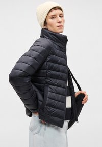 Melna pūkaina jaka ar augstu apkakli, iešūta tekstūra un ar rāvējslēdzēju priekšā. Valkāta ar krēmkrāsas adītu siltuma cepuri un svītrainu džemperi zemāk.