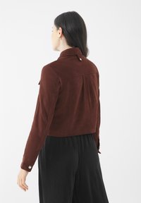 Hnědá semišová zkrácená bunda s límečkem, knoflíkem vzadu a plisovanými detaily. Nošena s volnými černými plisovanými kalhotami.