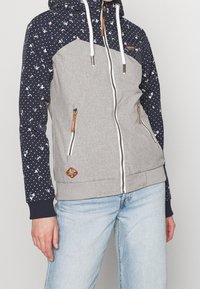 Grå zip-up hoodie med ett mönstrat navyhuva och ärmar, med blommönster och vita prickar. Inkluderar dragsko och logolapp.