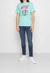 Camiseta de algodón turquesa con estampado gráfico, combinada con jeans ajustados azules y zapatillas blancas. Diseño sencillo con mangas cortas y cuello redondo.