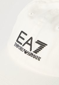 Șapcă albă din bumbac, cu logo EA7 brodat negru și textul „EMPORIO ARMANI”. Formă structurată cu orificii de ventilație.