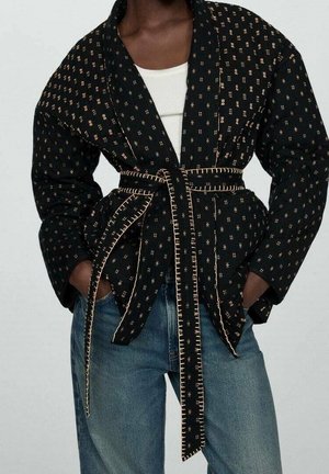 Schwarze, gemusterte Jacke mit Gürtel und hellbraunen Nähten, getragen über einem weißen Oberteil und blauen Jeans, Hände in den Taschen der Jacke.