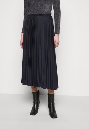 Kvinde iført sort plisseret midi-nederdel, glitrende langærmet top og sorte ankelstøvler, stående mod en ensfarvet baggrund.