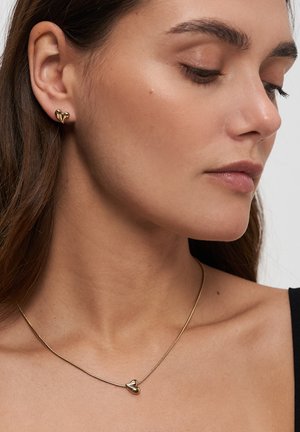 Frau, die goldene herzförmige Ohrstecker und eine passende goldene Herz-Anhängerhalskette trägt, mit neutralem Gesichtsausdruck.