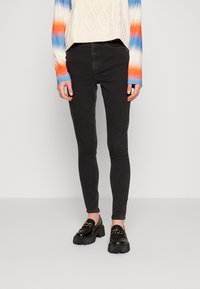 Jeans skinny negros con una textura suave, combinados con unos mocasines negros chunky con detalles de cadena dorada. La parte superior tiene un diseño degradado de azul y naranja.