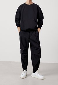 Felpa nera con tasca frontale, abbinata a pantaloni jogger neri coordinati con polsini elasticizzati e tasche laterali. Indossati con sneakers bianche.