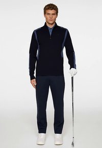 Marinblå tröja med ljusblå detaljer, dragkedjehals, kombinerad med marinblå byxor och vita sneakers. Golfklubba hålls i höger hand, vänster hand med handske.