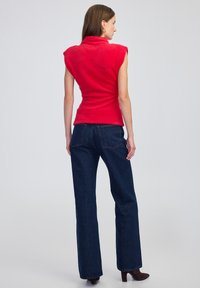 Žena stojící zády, oblečená v bezrukávovém červeném huňatém topu, tmavomodrých rovně střižených džínách a tmavých vysokých kotníkových botách před jednobarevným pozadím.