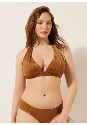 Frau mit welligem braunem Haar, die ein braunes Neckholder-Bikinitop und passende Höschen trägt, steht vor einem einfarbigen hellen Hintergrund.