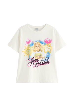 Nastoletnia dziewczyna uśmiecha się, ubrana w białą koszulkę z grafiką Zara Larsson, jasne dżinsowe szorty i brązowe klapki, stojąca w studio.