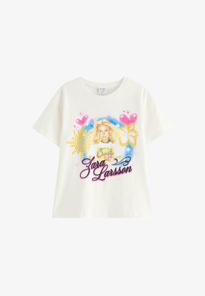 Nastoletnia dziewczyna uśmiecha się, ubrana w białą koszulkę z grafiką Zara Larsson, jasne dżinsowe szorty i brązowe klapki, stojąca w studio.
