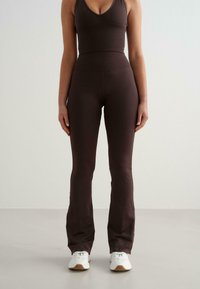 Femme debout portant un pantalon évasé ajusté marron foncé et un top sans manches assorti, avec des baskets blanches sur un fond clair.