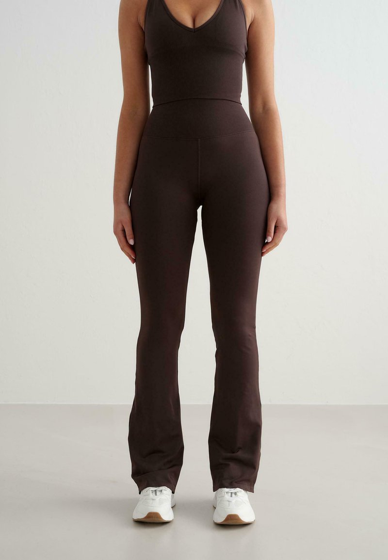 Femme debout portant un pantalon évasé ajusté marron foncé et un top sans manches assorti, avec des baskets blanches sur un fond clair.