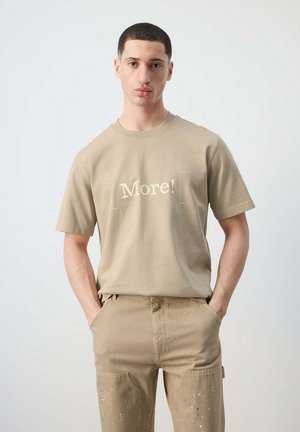 Joven mirando al frente con las manos en los bolsillos, vistiendo una camiseta beige con el texto "More!" y pantalones beige a juego.