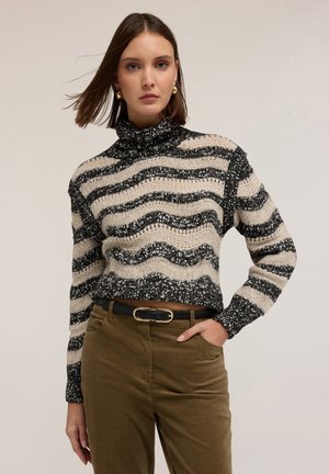 Suéter de punto con un patrón de zigzag en negro y crema, con un cuello alto. Combinado con pantalones verde oliva y un cinturón negro.