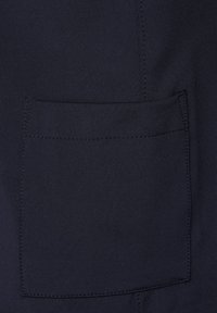 Námětový textil s hladkým povrchem obsahuje velkou náprsní kapsu obkreslenou pečlivým švem, umístěnou na jednolitém pozadí.