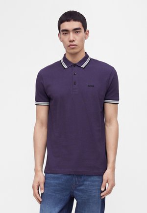 Giovane uomo che indossa una polo viola scuro con colletto e polsini a righe bianche, abbinata a jeans blu, in piedi su sfondo bianco.